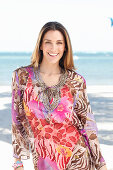 A brunette woman by the sea wearing a colourful beach dress