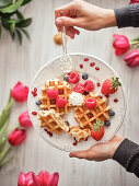 Hands holding dish with delicious waffles and fresh berries on blurred background