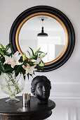 Bust and vase of lilies on antique table below round mirror