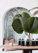 Decorative glass bottles and green palm fan leaves in vase on table in front of alcove