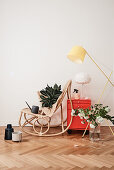 Rattan-Schaukelstuhl mit Zimmerpflanze und Gießkanne, rotes Schränkchen und Stehlampe