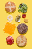 Different ingredients of a cheese burger wrapped in plastic on yellow background
