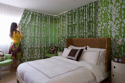 Doppelbett mit Rattan-Kopfteil, Nachttisch und grüner Stuhl im Schlafzimmer, Frau zieht Vorhang zu