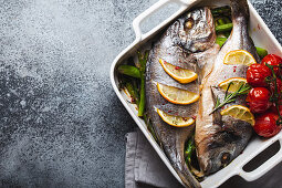 Baked fish dorado with green asparagus and tomatoes in white ceramic baking pan on gray rustic concrete background