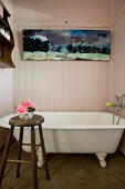 Vintage bathtub below painting in wood-clad bathroom with bowl of flowers on stool