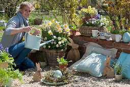 Woman pours basket with daffodils and horned violets, Easter pebble terrace with wooden Easter bunnies and wreath of branches