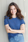 A young brunette woman in front of a grey wooden wall wearing a blue t-shirt