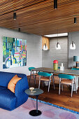 Blue sofa and side table, behind it dining table with chairs in an open living room with a wooden ceiling and concrete-look walls