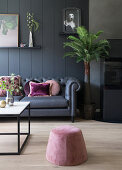 A dark sofa with pink cushions in front of a dark wall with a pouffe and a coffee table in the foreground