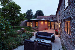 Blick über Terrasse mit Outdoor-Möbeln auf beleuchteten Anbau aus Ziegelsteinen und auf bestehendes Bauernhaus
