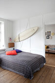 A double bed with a bedspread in a bedroom with rustic floorboards and surfboard on a white wall