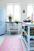 Light blue, open shelf with colorful dishes in the kitchen-living room, in the background a refrigerator next to the window