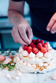 A two-tier meringue cake being decorated with raspberries