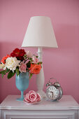 Rosenstrauß, Tischleuchte und nostalgischer Wecker auf Nachtschrank vor rosa Wand
