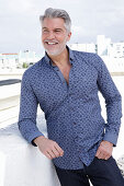 A grey-haired man on a terrace wearing a blue shirt and jeans