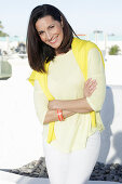 A mature brunette woman wearing a yellow top and white trousers with a jumper over her shoulders