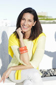 A mature brunette woman wearing a yellow top and white trousers with a jumper over her shoulders