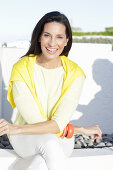 A mature brunette woman wearing a yellow top and white trousers with a jumper over her shoulders