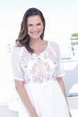 A long-haired woman wearing a white summer dress with colourful embroidery detail