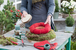 Homemade moss wreath cake with candle: woman puts red felt wreath around a clay pot and wooden disc over it
