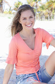 A long-haired woman wearing a short-sleeved salmon-coloured top and denim shorts