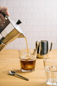 Pouring coffee from coffeemaker into stylish glass with ice on wooden counter in cafe