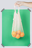 Hand carrying net bag with ripe oranges against green rectangle during zero waste shopping