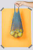 Hand carrying net bag with ripe apples against orange rectangle during zero waste shopping