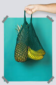 Hand carrying net bag with ripe pineapple and bananas against blue turquoise rectangle during zero waste shopping