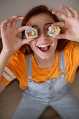 Young female keeping tasty sushi near eyes while having lunch at home