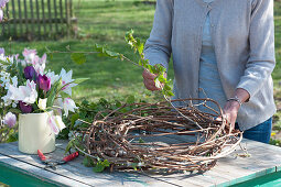 Frau windet Kranz aus Ranken und Zweigen, Krug mit Tulpen