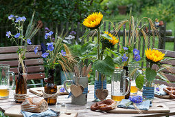 Table decoration for a Bavarian snack with cranesbill and ears of cereal in beer bottles, sunflowers, cutlery in a beer mug with a wooden heart as decoration
