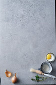 A grey surface with an onion, flour, egg, herbs and a brush