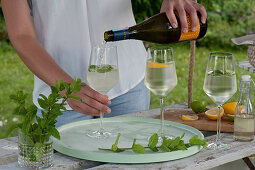 Frau füllt Gläser mit Prosecco, Strauß aus frischer Minze, Orangen, Limetten für Hugo