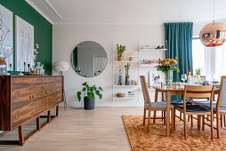 A sideboard in front of a green wall, a round mirror on the wall and an open shelf in a dining room