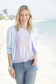 A blonde woman by the sea wearing a purple t-shirt and a blue-and-white striped shirt