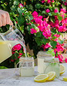 Pouring fresh homemade lemonade from pitcher into glass jars placed on table in blooming summer garden