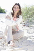 A long-haired woman sitting in the sand on the beach wearing a light jumper and trousers