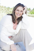 A long-haired woman sitting in the sand on the beach wearing a light jumper and trousers