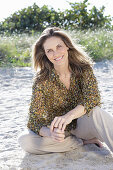 A long-haired woman sitting in the sand wearing a floral shirt and light trousers