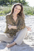 A long-haired woman sitting in the sand wearing a floral shirt and light trousers