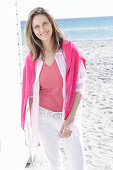 A long-haired woman on the beach wearing a long blouse with a pink jumper over her shoulders