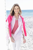 A long-haired woman on the beach wearing a long blouse with a pink jumper over her shoulders