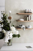 Wooden shelves with dishes on the wall, bouquet of white flowers on table