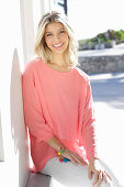 A young blonde woman wearing a salmon-pink blouse and white trousers