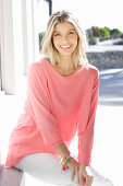 A young blonde woman wearing a salmon-pink blouse and white trousers