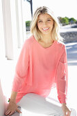 A young blonde woman wearing a salmon-pink blouse and white trousers