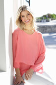 A young blonde woman wearing a salmon-pink blouse and white trousers