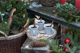 Mug with hot mulled cider and cinnamon sticks on wooden disc, basket with fir branches, and old wooden skates