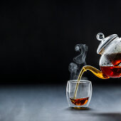 Dampfender schwarzer Tee wird aus Teekanne in Glasbecher gegosssen